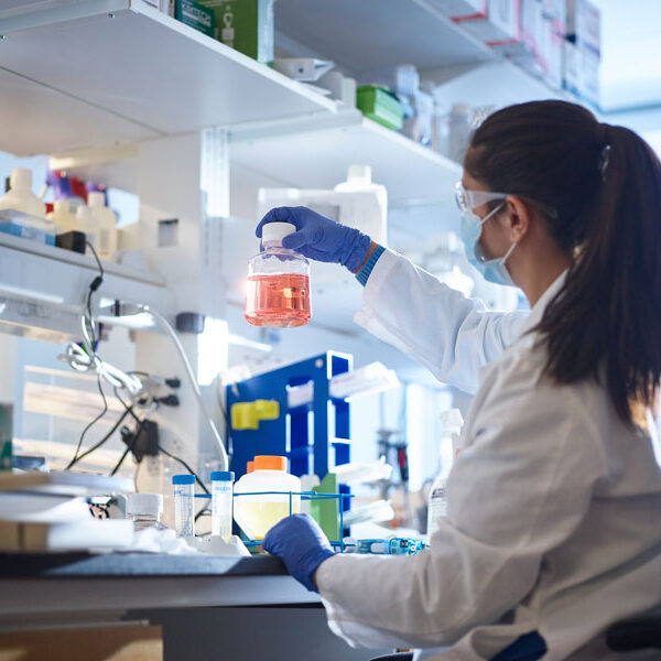 A female individual donning medical gloves and a mask is seen showcasing a bottle containing fluid.